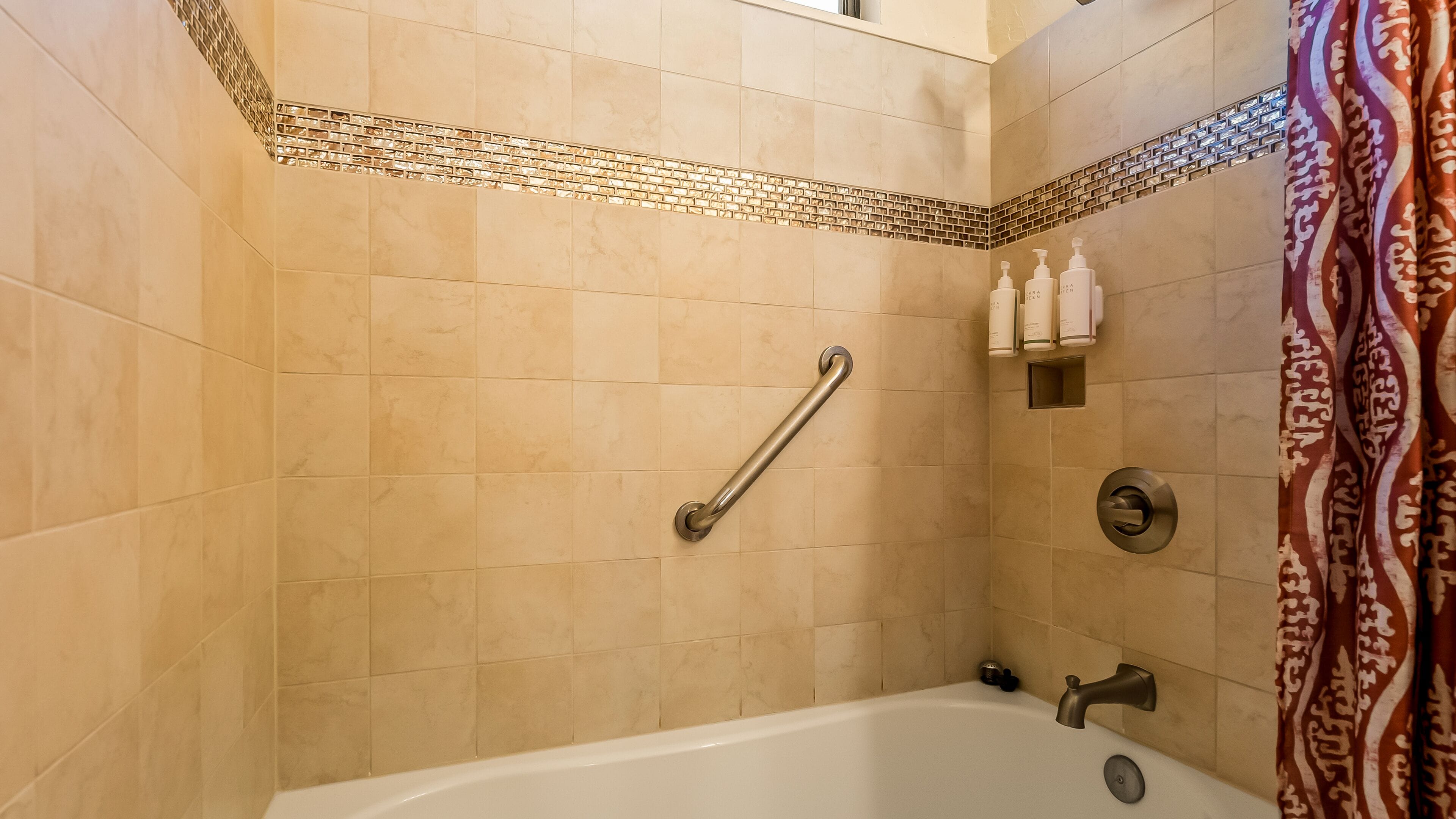 A bathroom with beige tiles, a grab bar, a showerhead, and a toilet paper holder.