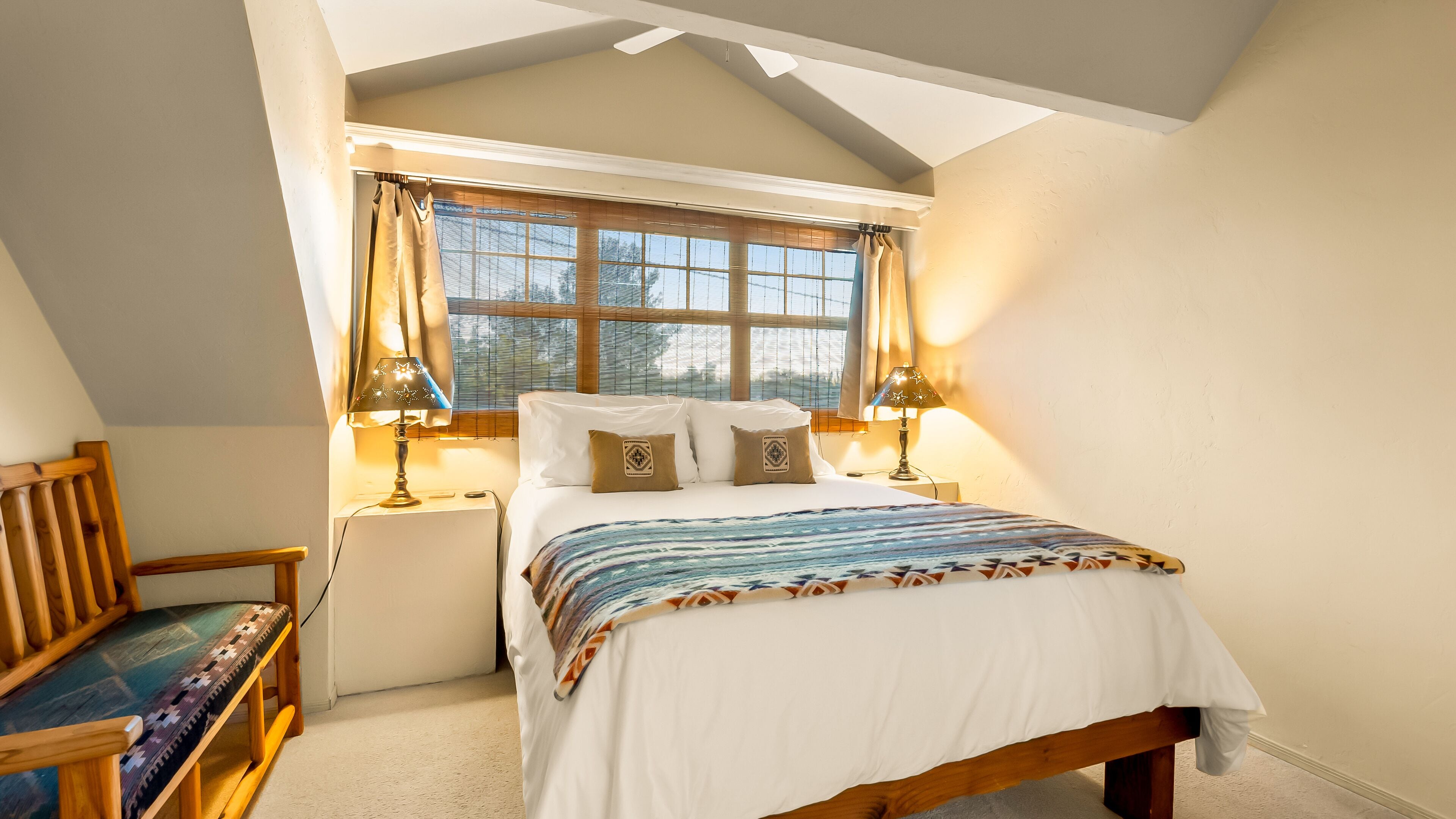 A bedroom with a sloped ceiling, a bed with a patterned bedspread, two bedside lamps, and a window with curtains.
