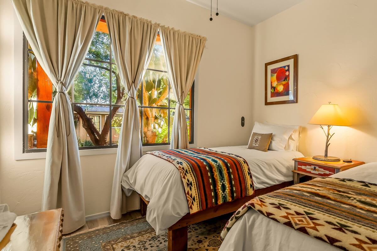 A hotel room with two beds, a window with curtains, a bedside lamp, and a painting on the wall.