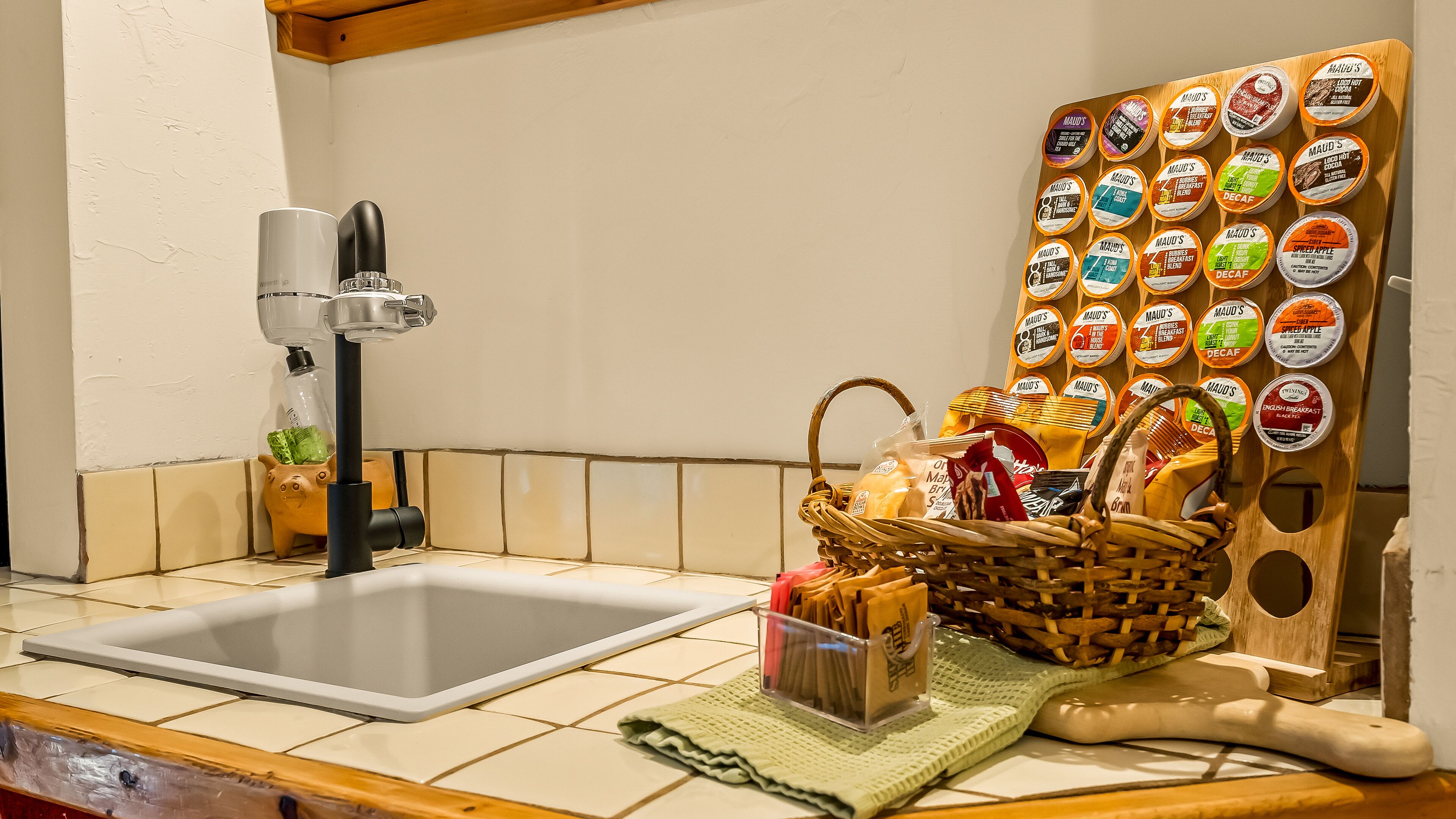A kitchen with a sink, a faucet, a wooden board with food containers, a basket of bread, and a rolling pin.