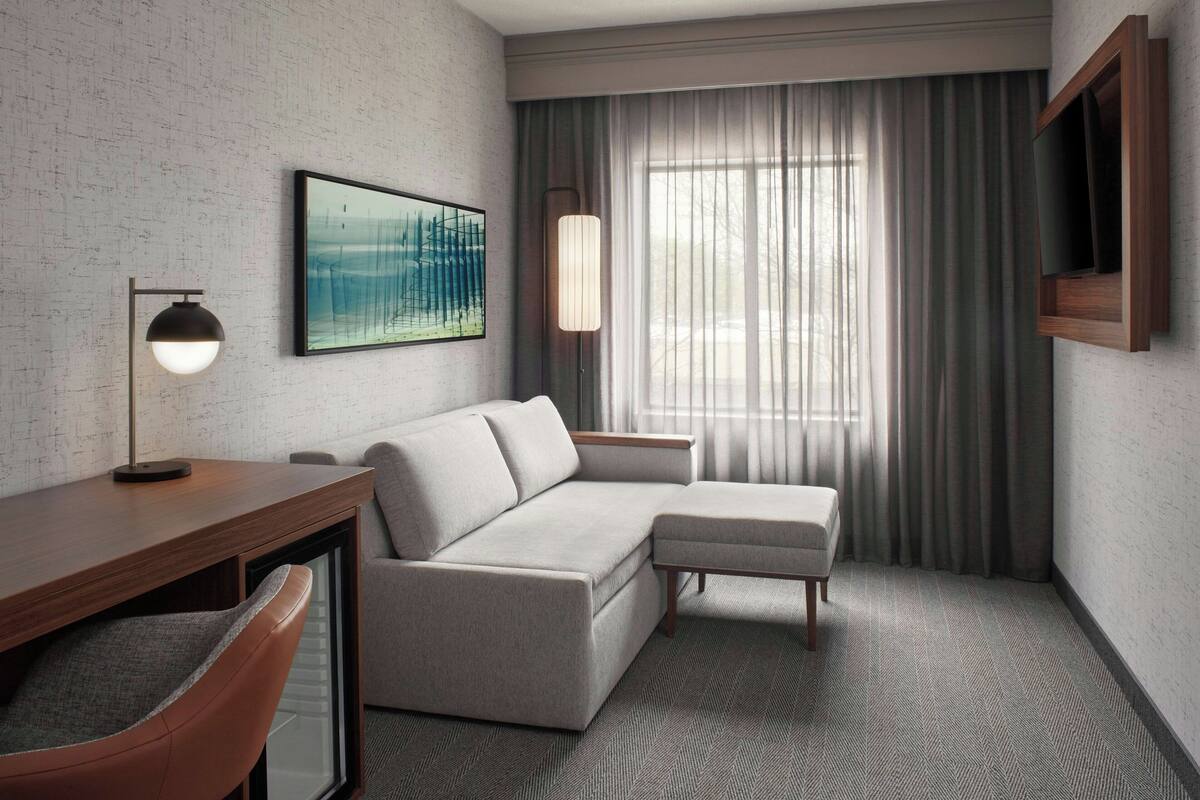 A modern hotel room with a sofa, a desk, a chair, a TV mounted on the wall, and a large window with curtains.