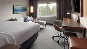 Une chambre d’hôtel avec un grand lit, un bureau, une chaise, une télévision et une fenêtre donnant sur des arbres.