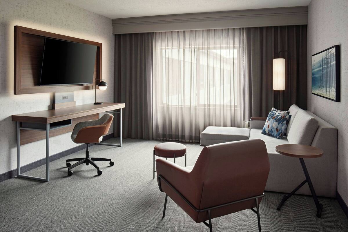 A modern hotel room with a flat-screen TV mounted on the wall, a wooden desk, a leather armchair, a round coffee table, a sofa, and a large window with sheer curtains.