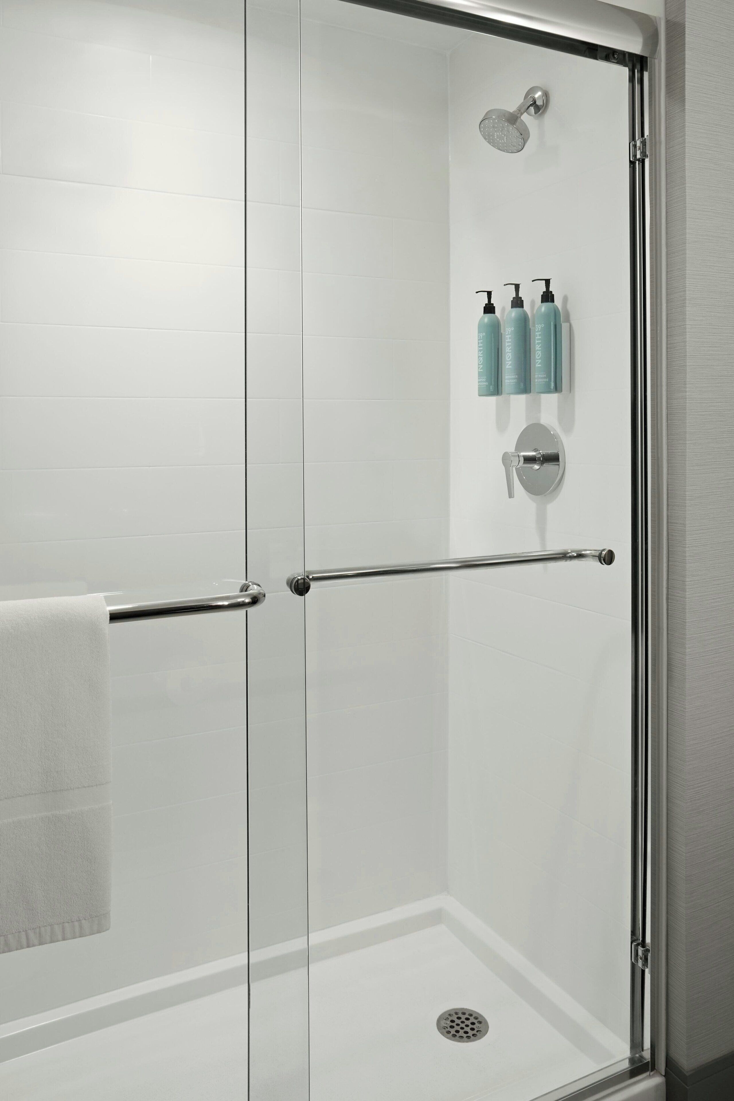 A glass-enclosed shower with a metal grab bar, a white tiled wall, and three bottles of shampoo on the wall.