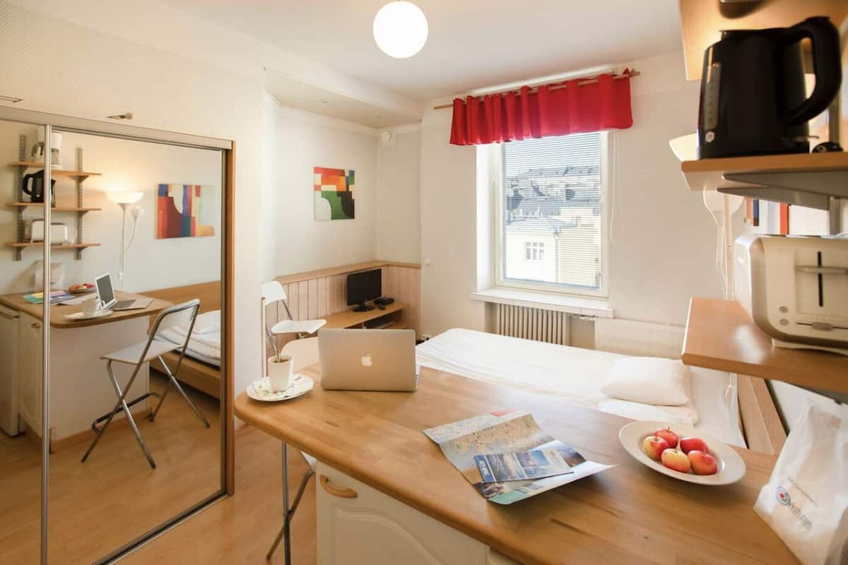 A compact hotel room with a wooden desk, a laptop, a bed, a window with a red curtain, and a radiator.