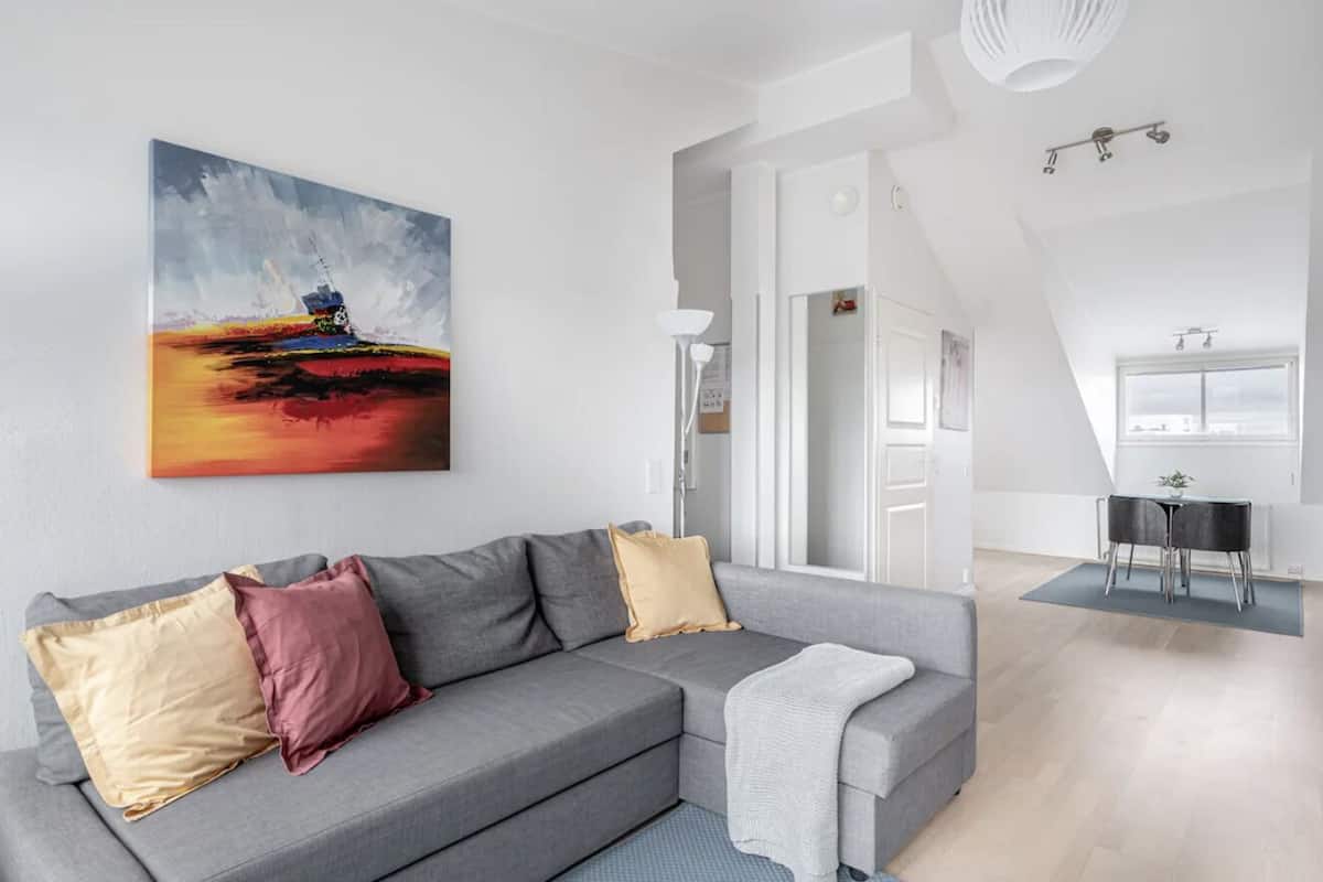 A modern living room with a grey sofa, a painting, and a dining area with a table and chairs.