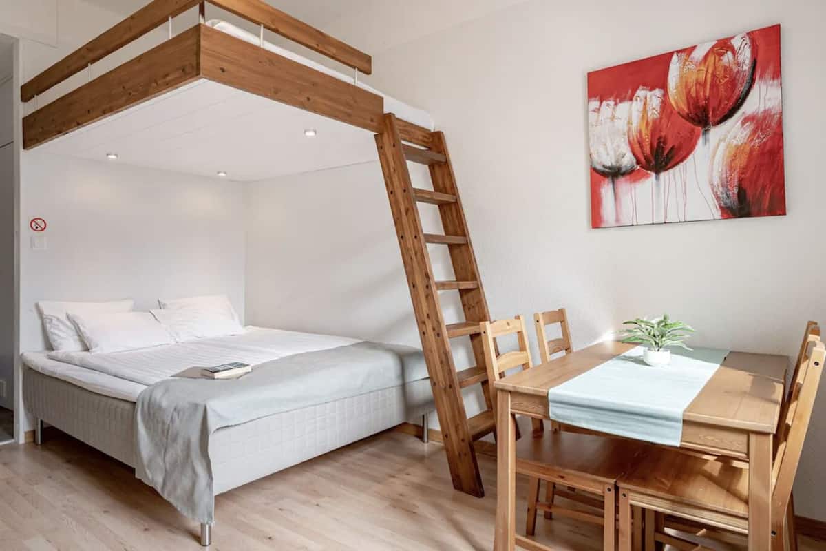 A bedroom with a bunk bed, wooden ladder, a dining table with chairs, and a painting on the wall.