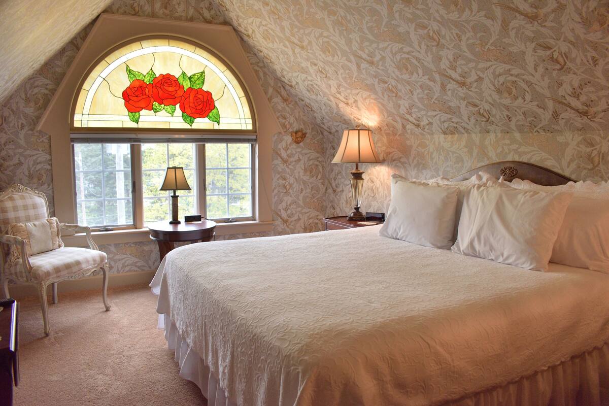 A bedroom with a large bed, a chair, a nightstand, a lamp, a window with a stained glass rose design, and patterned wallpaper.