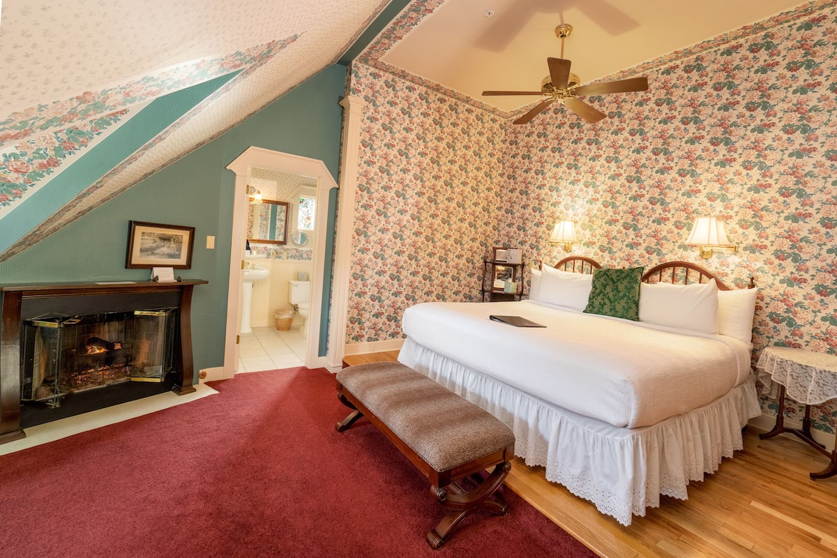 A bedroom with a bed, a bench, a fireplace, and a ceiling fan.