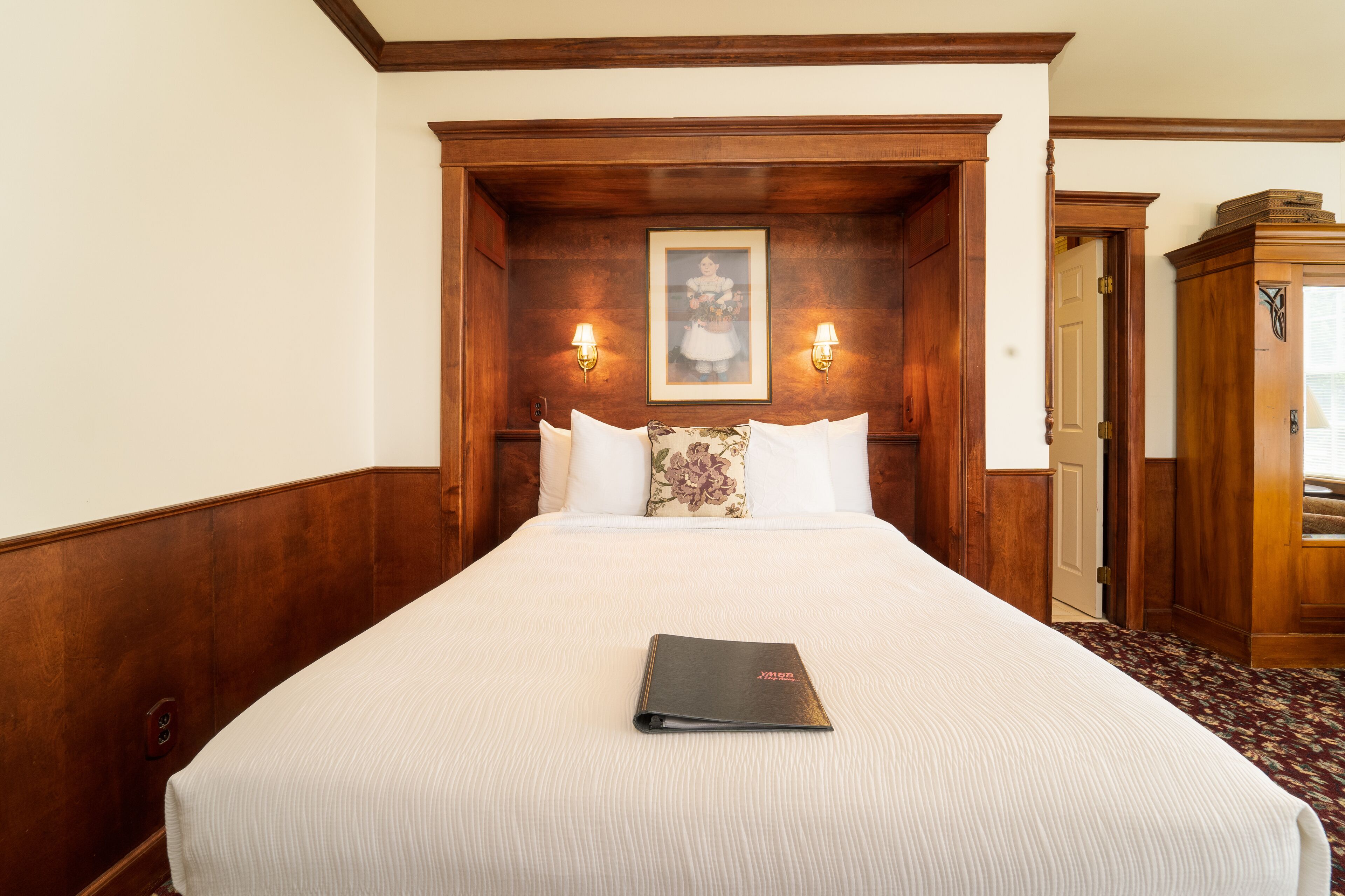 Habitación de hotel con una cama grande, cabecera de madera, dos lámparas fijadas a la pared y un cuadro enmarcado sobre la cama.