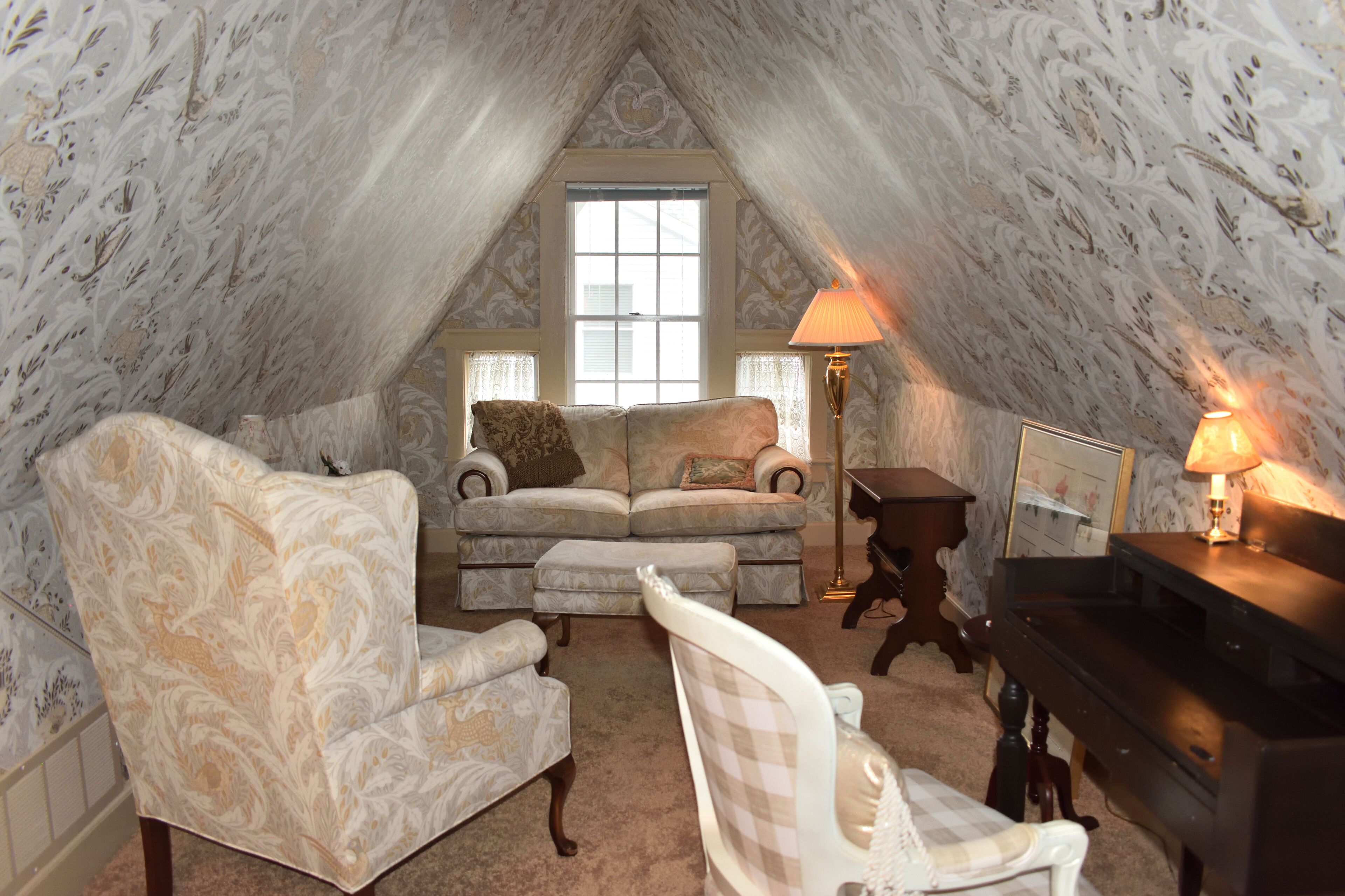 A cozy attic room with a large window, a piano, and a sofa set.