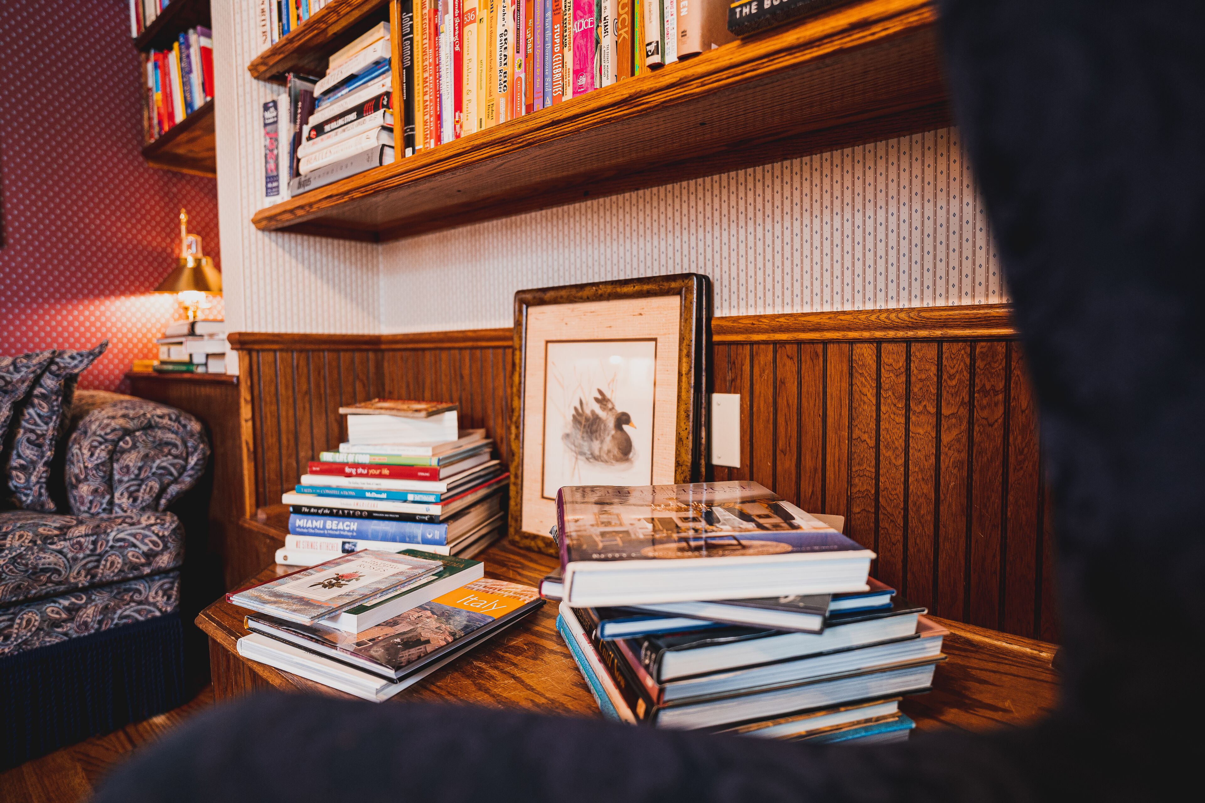 A cozy reading nook with a wooden bookshelf, a patterned armchair, and a small table with books and a framed picture.