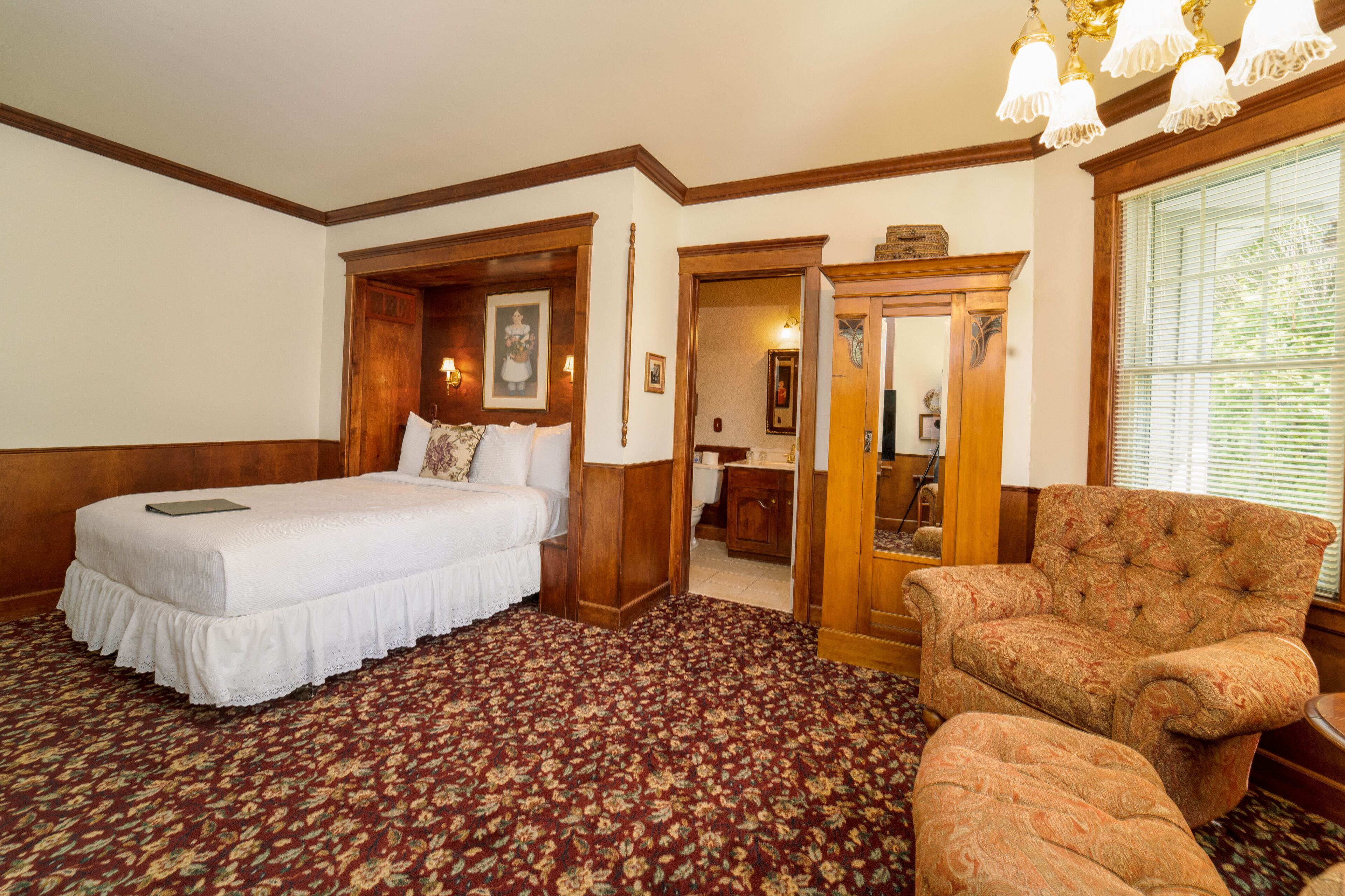 A hotel room with a large bed, a chair, a wooden wardrobe, and a mirror.