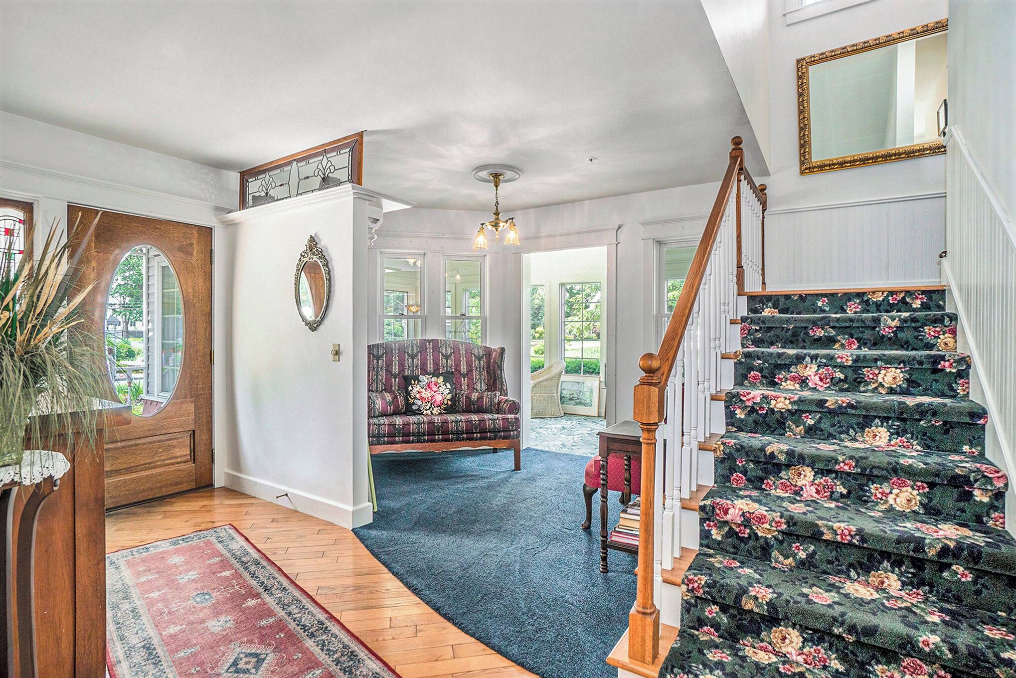 Un magnifique escalier recouvert d’une moquette à motifs floraux, une porte en bois ornée d’un miroir et un salon chaleureux avec un canapé et un lustre.