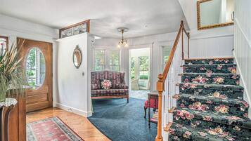 Un magnifique escalier recouvert d’une moquette à motifs floraux, une porte en bois ornée d’un miroir et un salon chaleureux avec un canapé et un lustre.