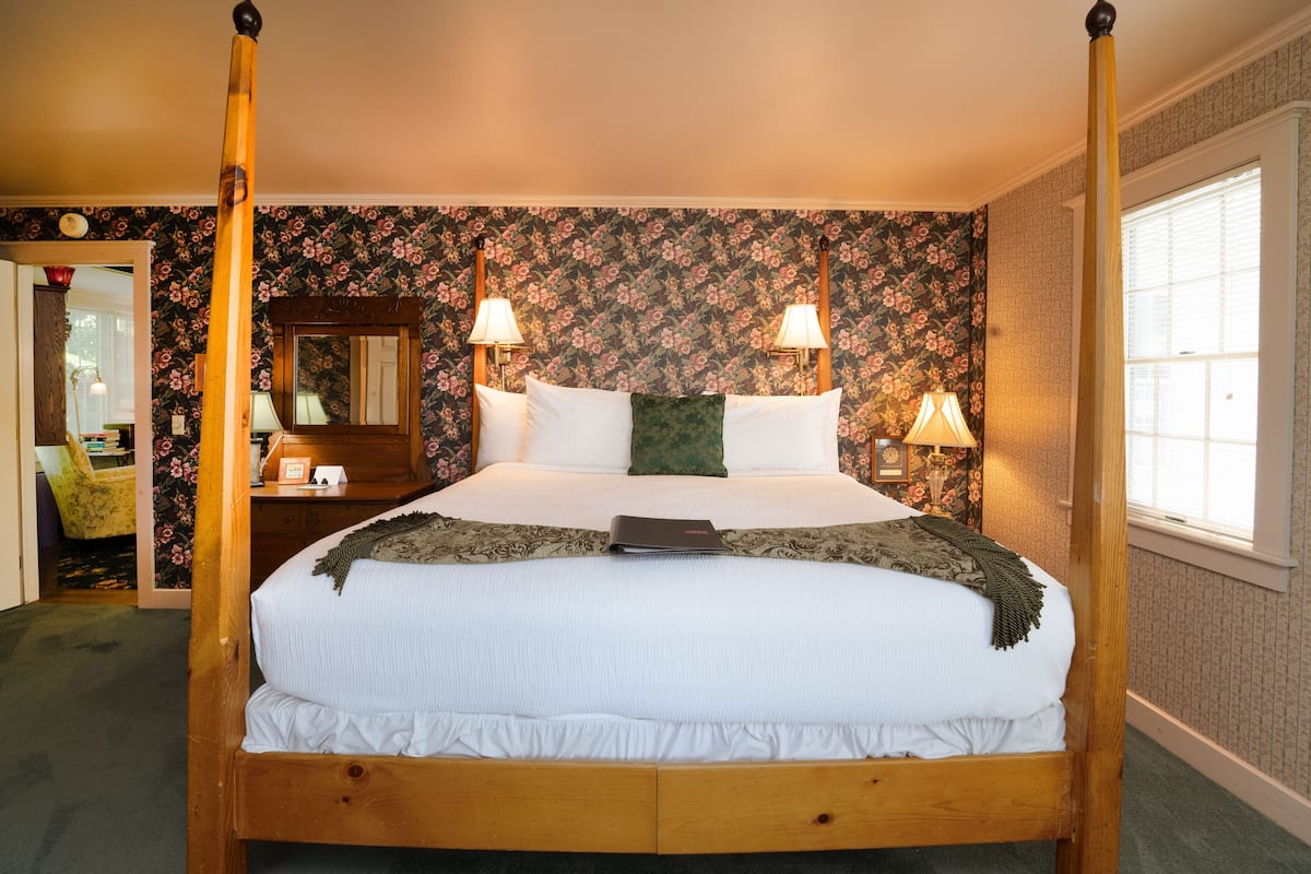 A four-poster bed with white bedding, two wall-mounted lamps, and a floral wallpaper pattern.