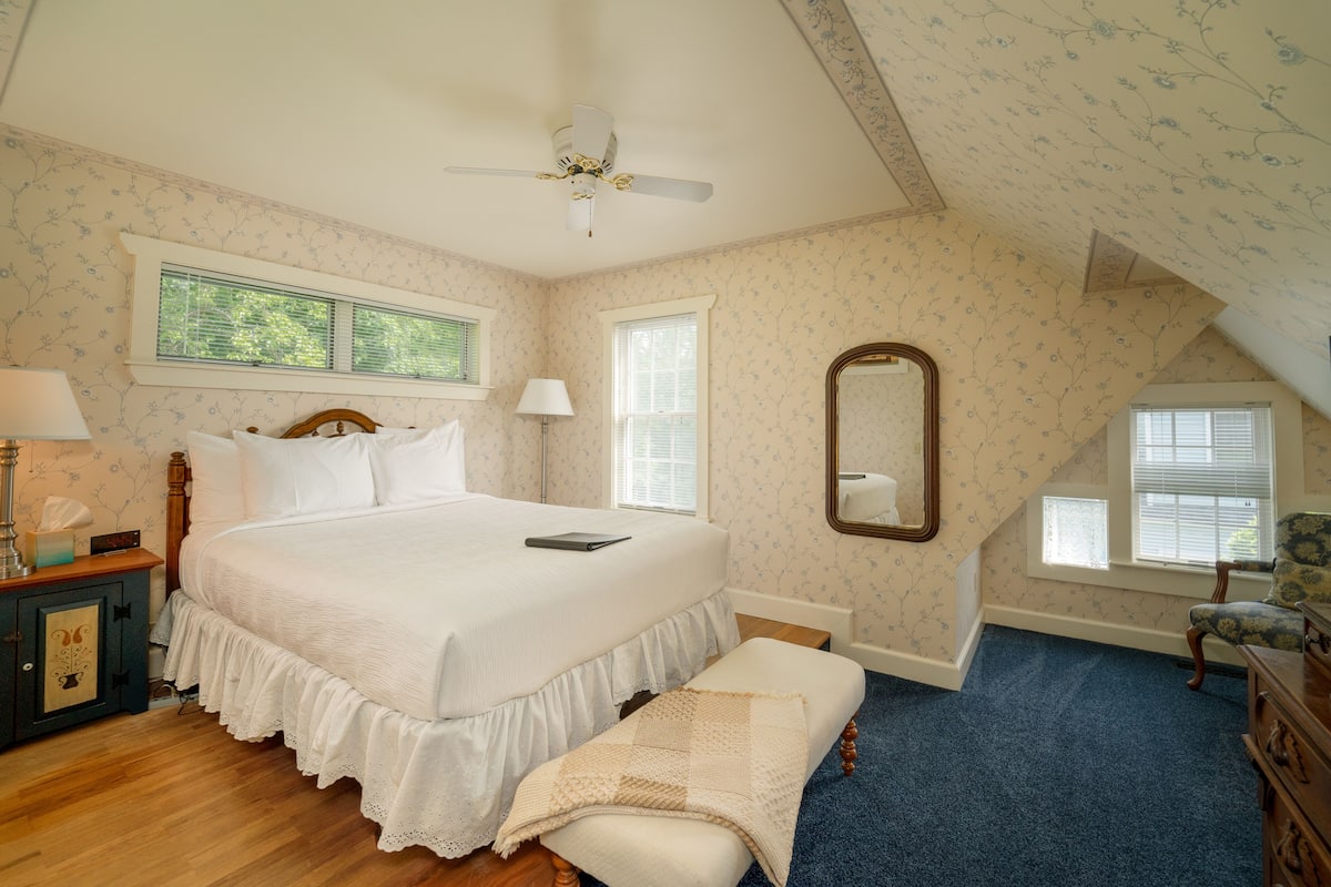 A bedroom with a large bed, a ceiling fan, two windows, a mirror, and a blue carpet.