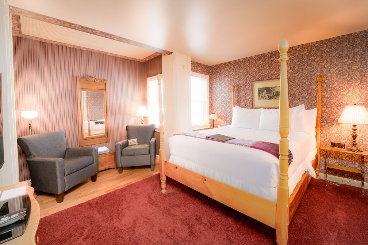 A bedroom with a four-poster bed, two armchairs, a wooden nightstand, and a red carpet.