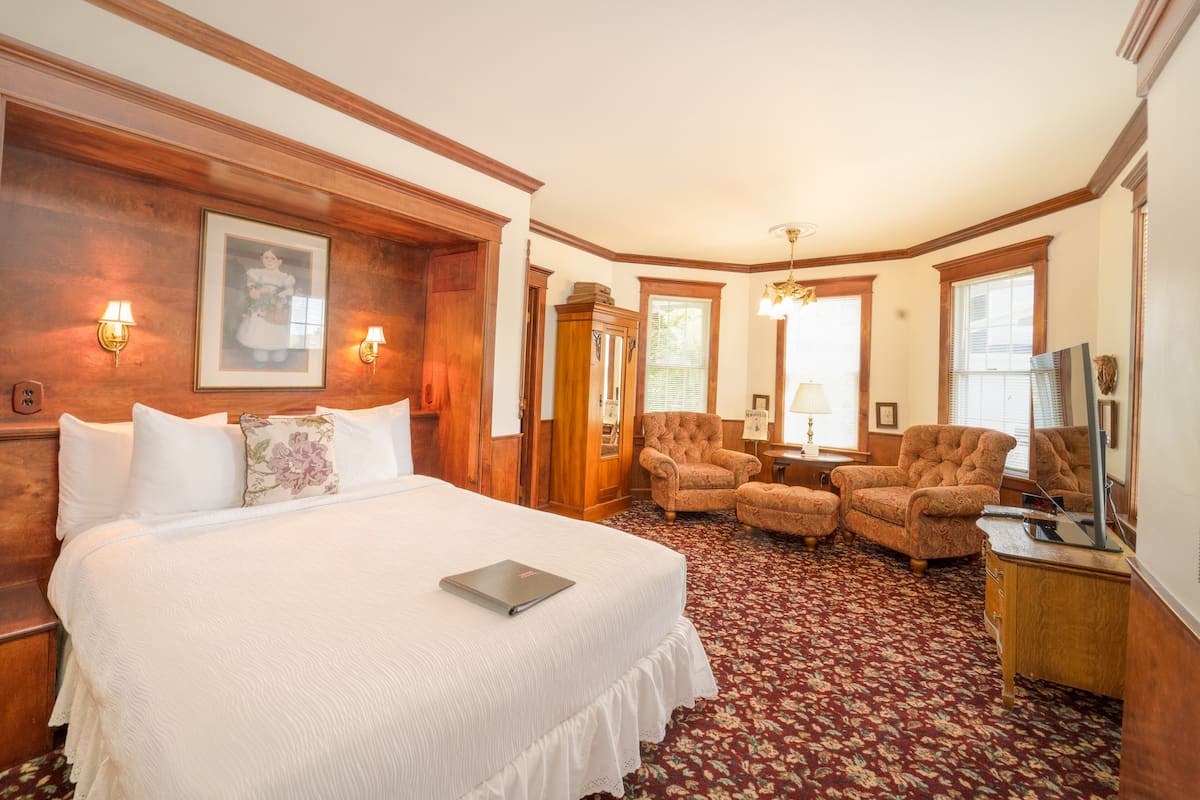 A hotel room with a large bed, two armchairs, a wooden wardrobe, and a desk with a lamp.