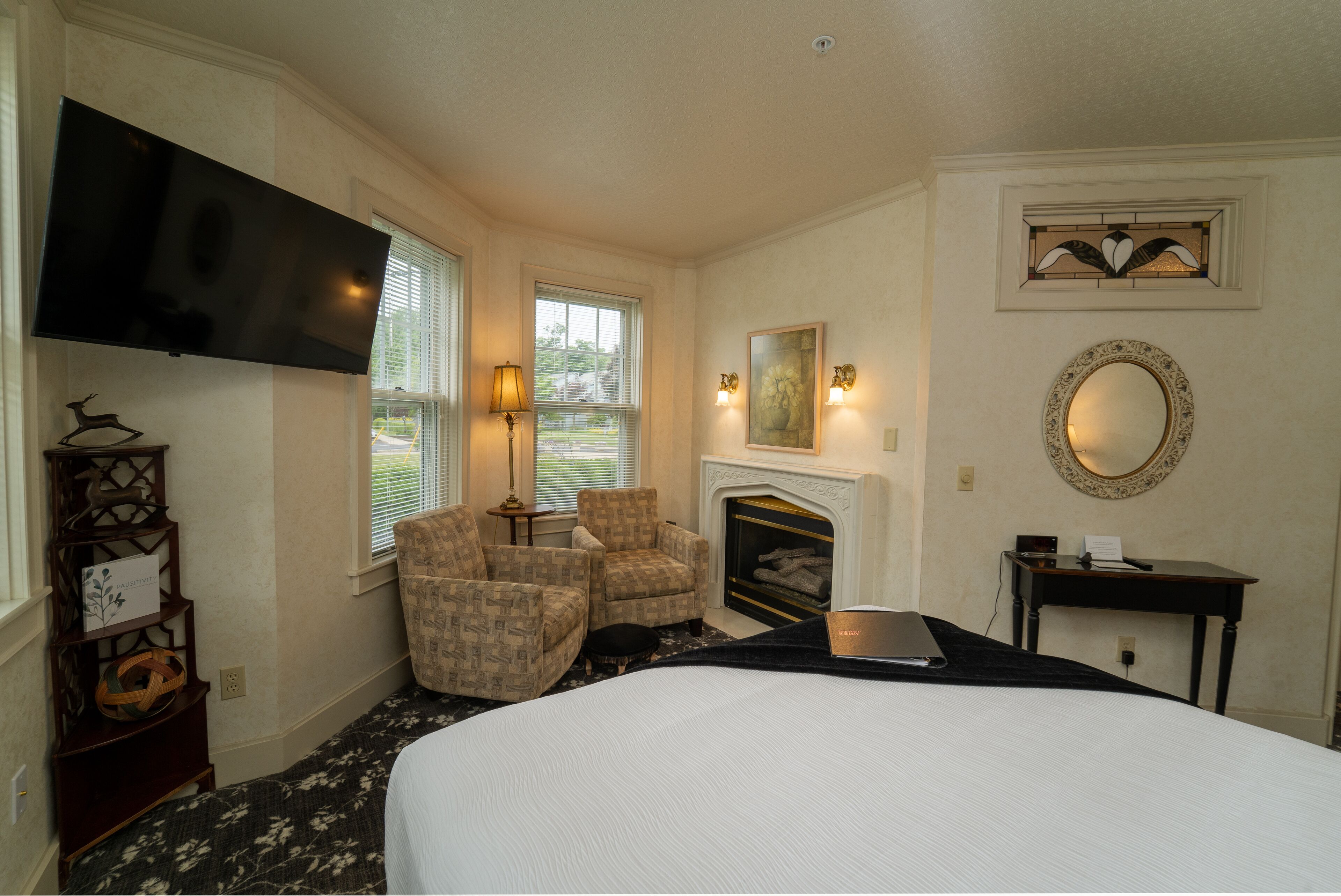 A hotel room with a large bed, two armchairs, a fireplace, a TV, and a desk with a mirror.