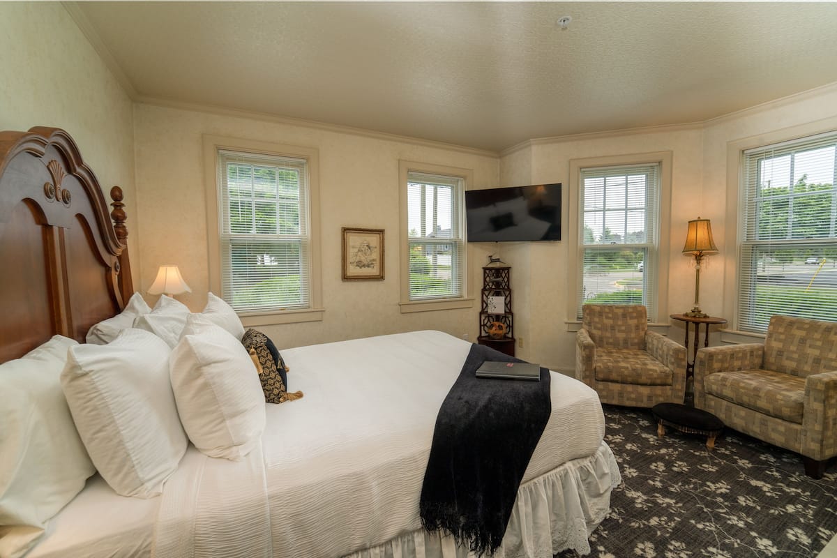 A hotel room with a large bed, two armchairs, a television, and a window with blinds.
