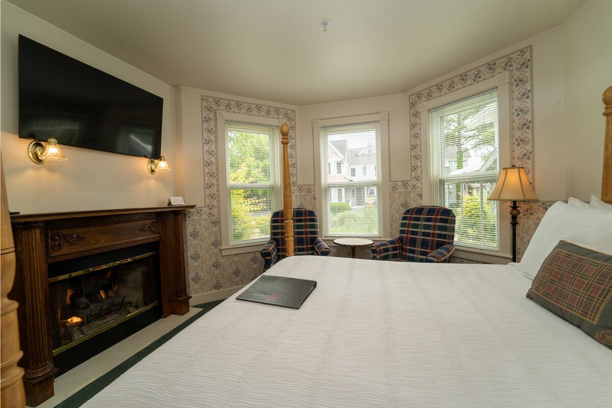 A hotel room with a large bed, a fireplace, two plaid chairs, a small table, and a TV mounted on the wall.