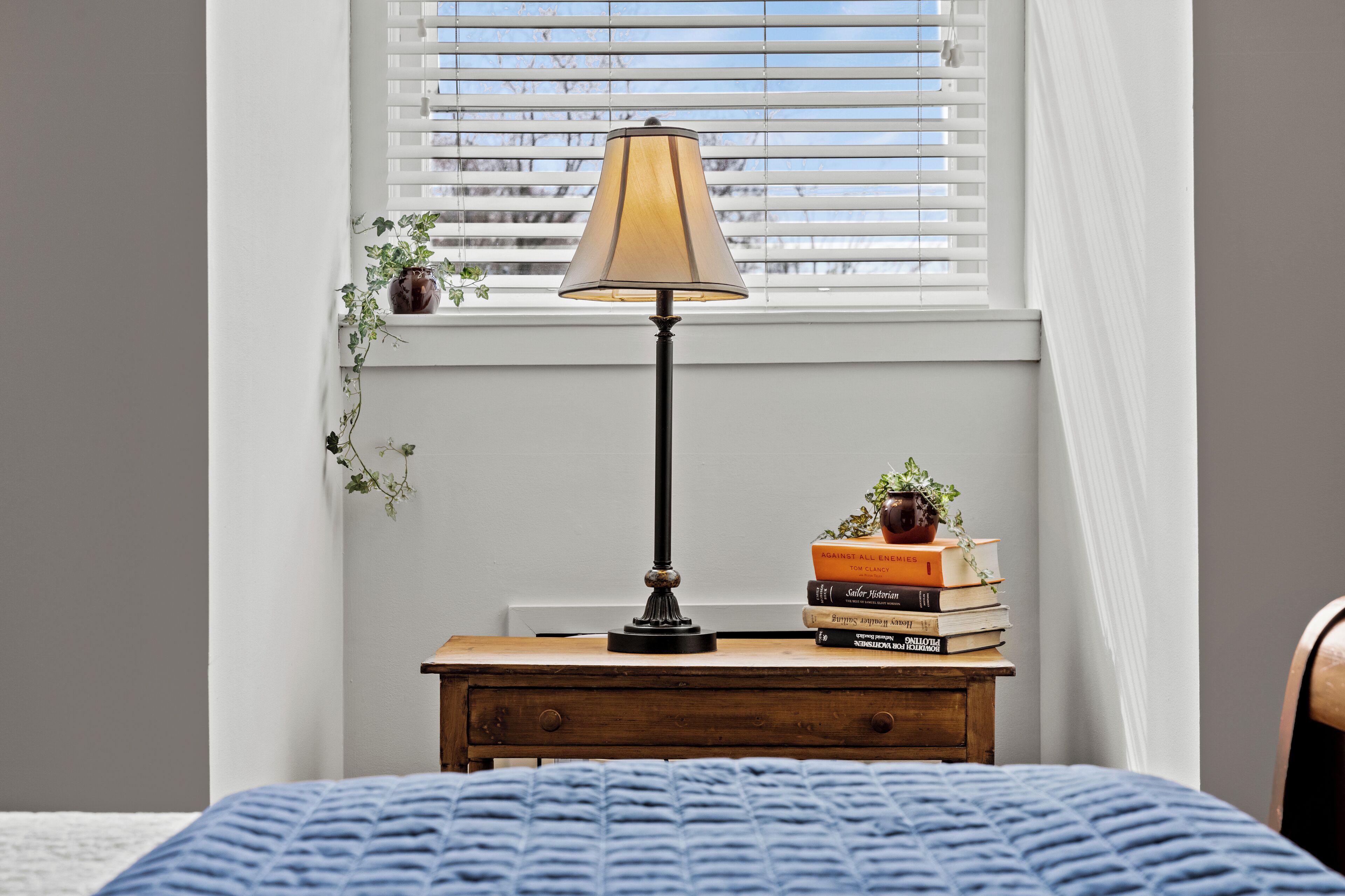 A bedroom with a bed, a bedside table with a lamp, a window with blinds, and a small plant on the window sill.