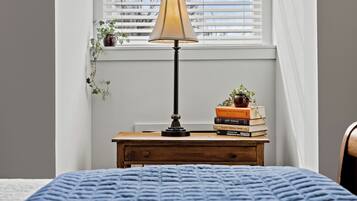 A bedroom with a bed, a bedside table with a lamp, a window with blinds, and a small plant on the window sill.