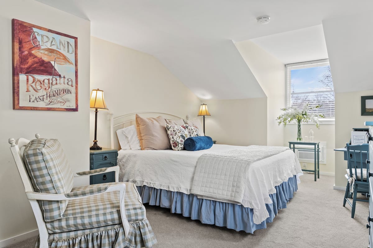A bedroom with a large bed, two bedside tables with lamps, a chair, a desk, and a window with a view of the sky.