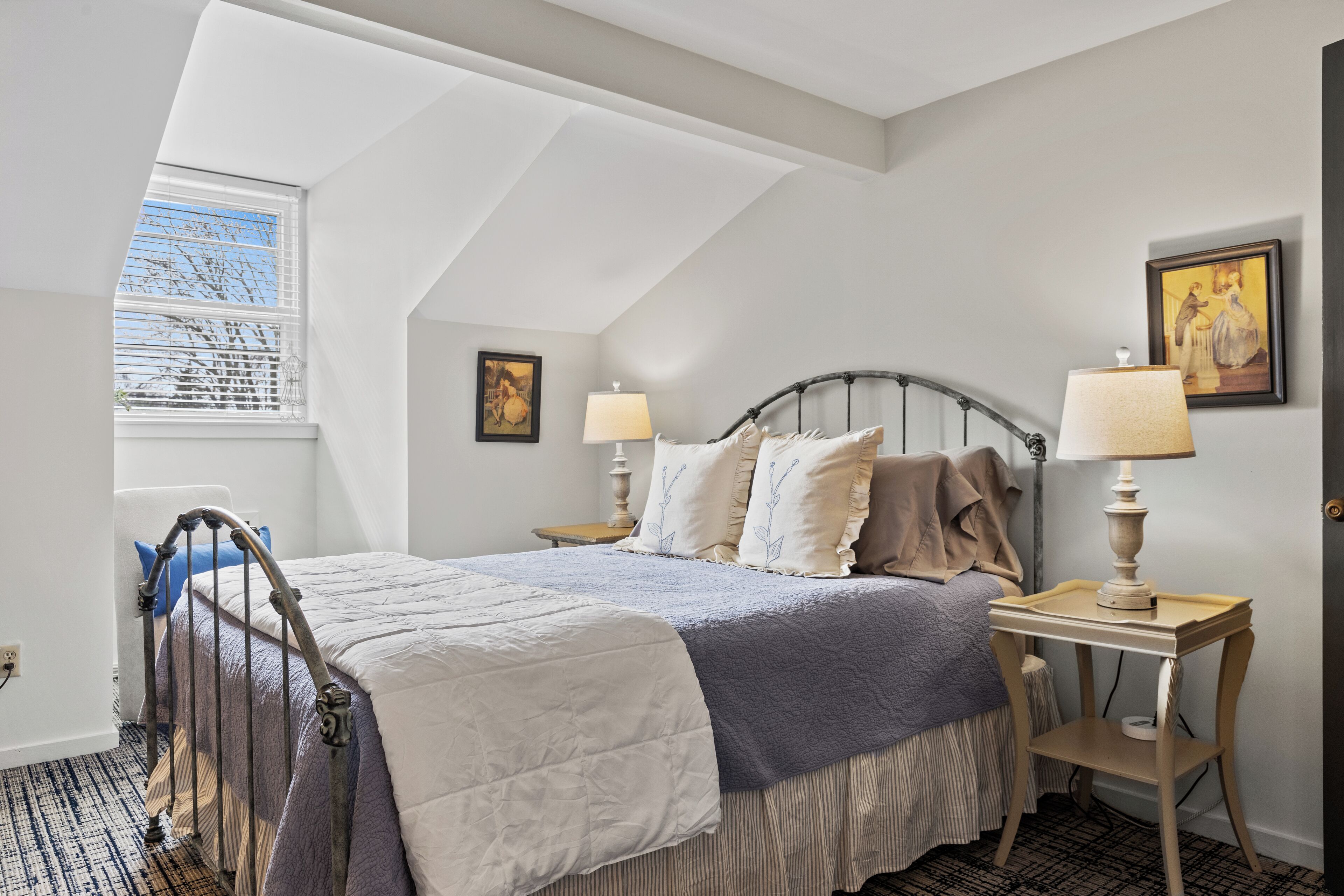 Une chambre à coucher avec un lit, deux tables de chevet avec des lampes, une fenêtre avec des stores et un tableau au mur.