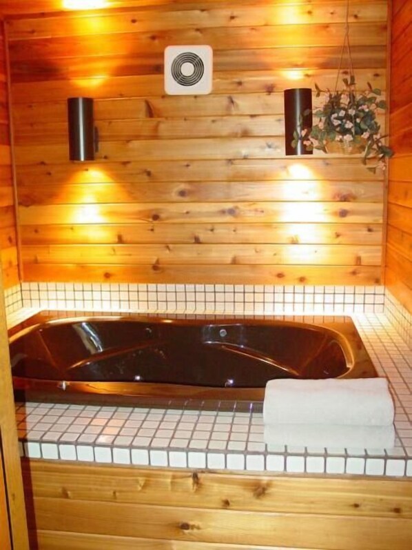 A wooden cabin interior with a wooden wall, a tiled floor, a black bathtub, a white towel, and a small plant. - Woodland Motor Lodge (Grayling)