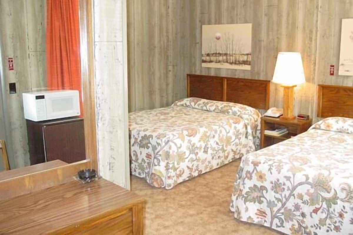 A hotel room with two beds, a wooden headboard, a bedside lamp, a microwave, and a small table.