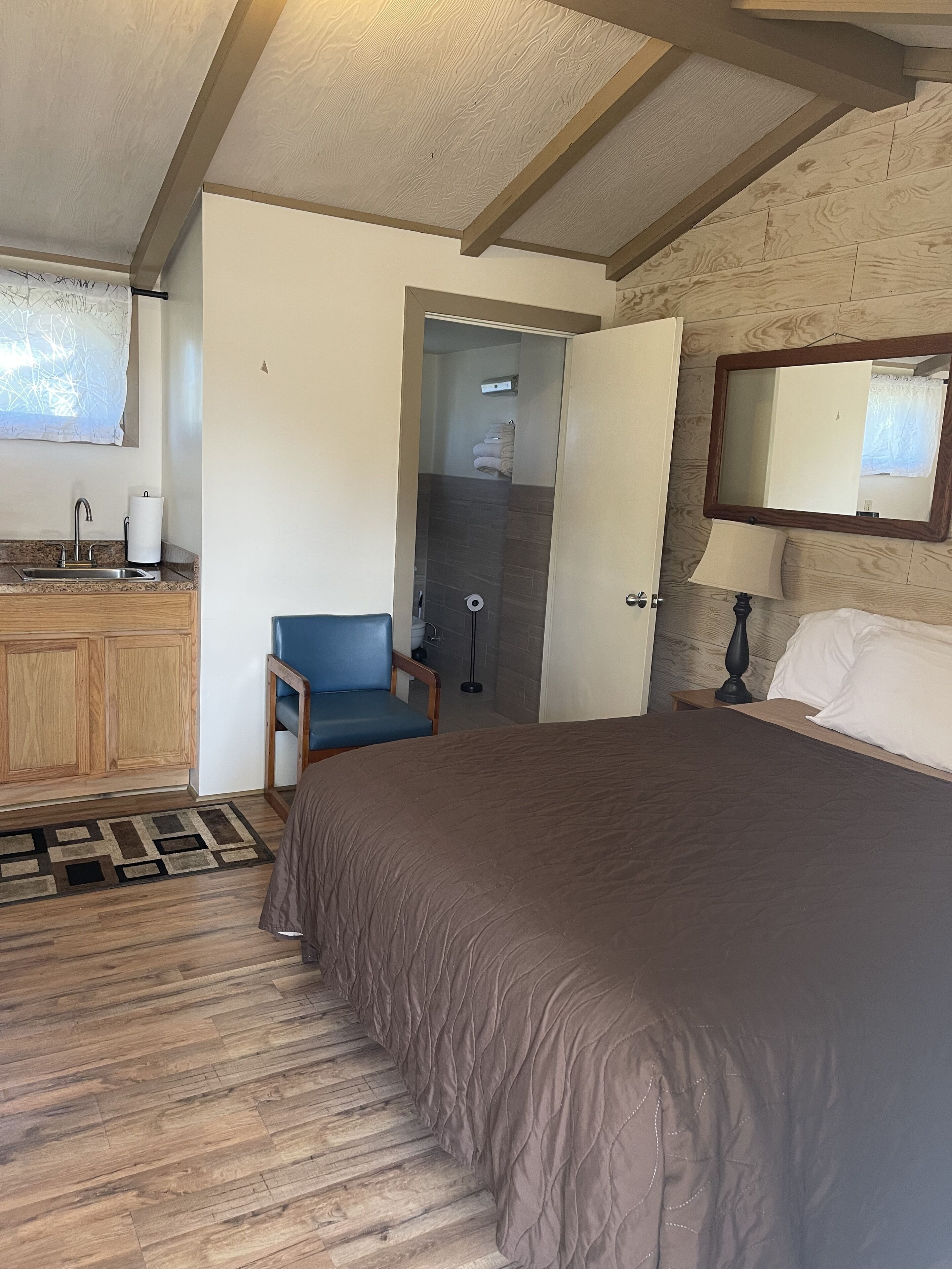 A bedroom with a large bed, a wooden dresser, a chair, a bedside table with a lamp, and a mirror on the wall.