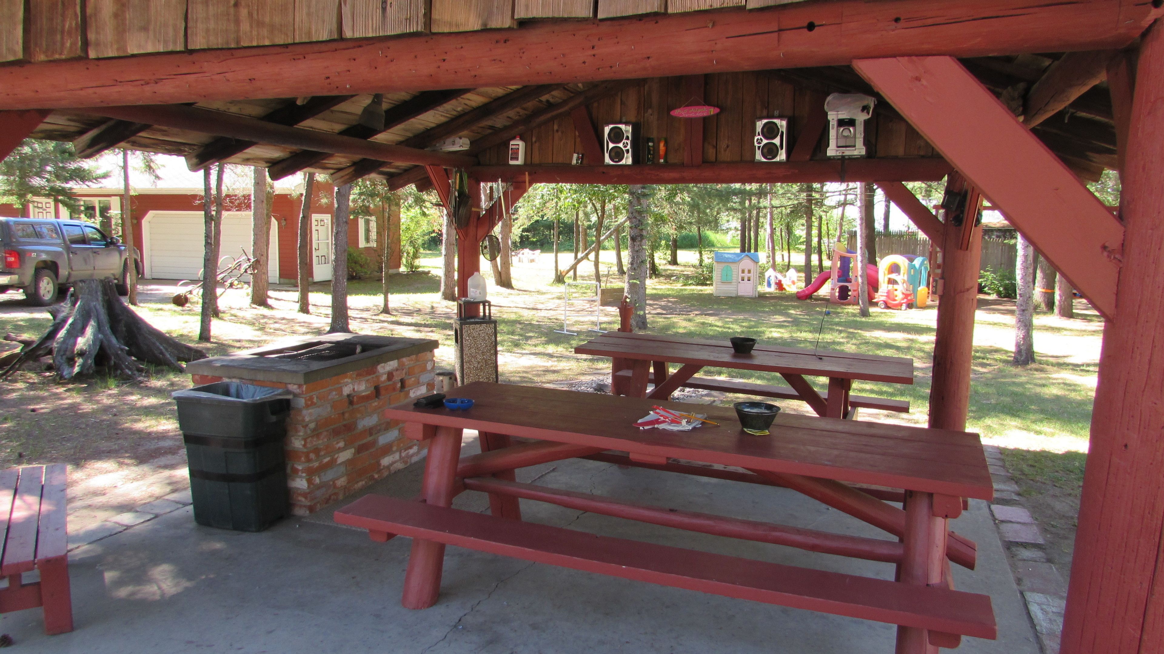 A wooden picnic shelter with a brick barbecue, picnic tables, and a trash bin.