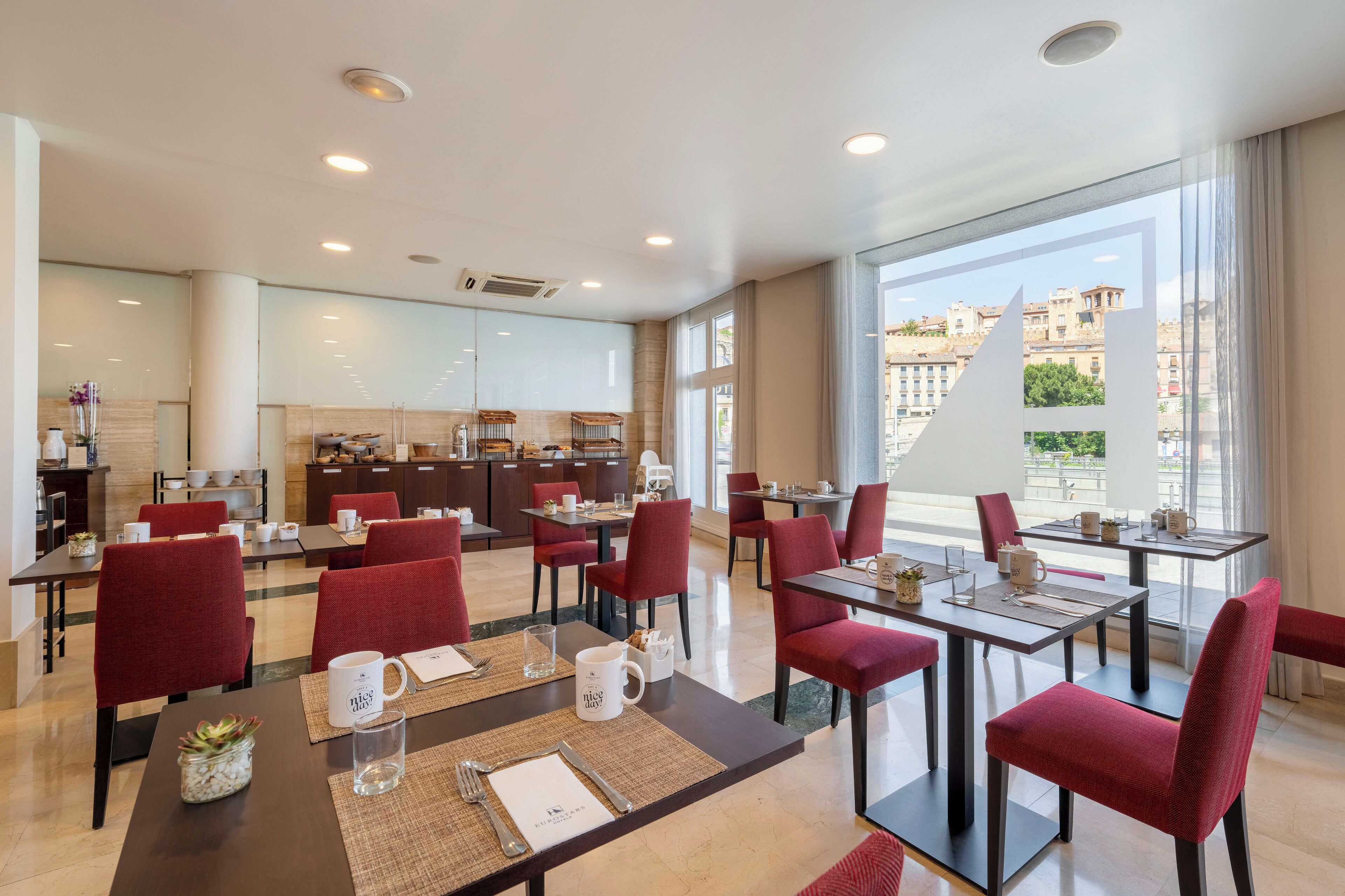Un comedor de hotel moderno con sillas tapizadas rojas, una isla central con electrodomésticos de cocina y ventanas amplias con vistas a la ciudad.