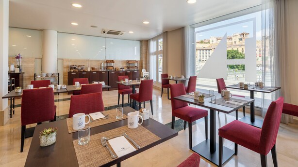 Un espace de restauration moderne dans un hôtel, avec des chaises à revêtement rouge, un îlot central doté d’appareils de cuisine et de grandes fenêtres offrant une vue sur la ville.