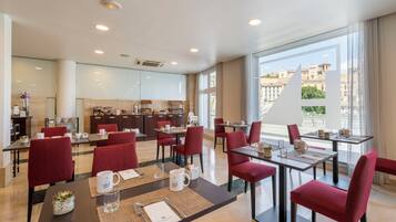 Un comedor de hotel moderno con sillas tapizadas rojas, una isla central con electrodomésticos de cocina y ventanas amplias con vistas a la ciudad.