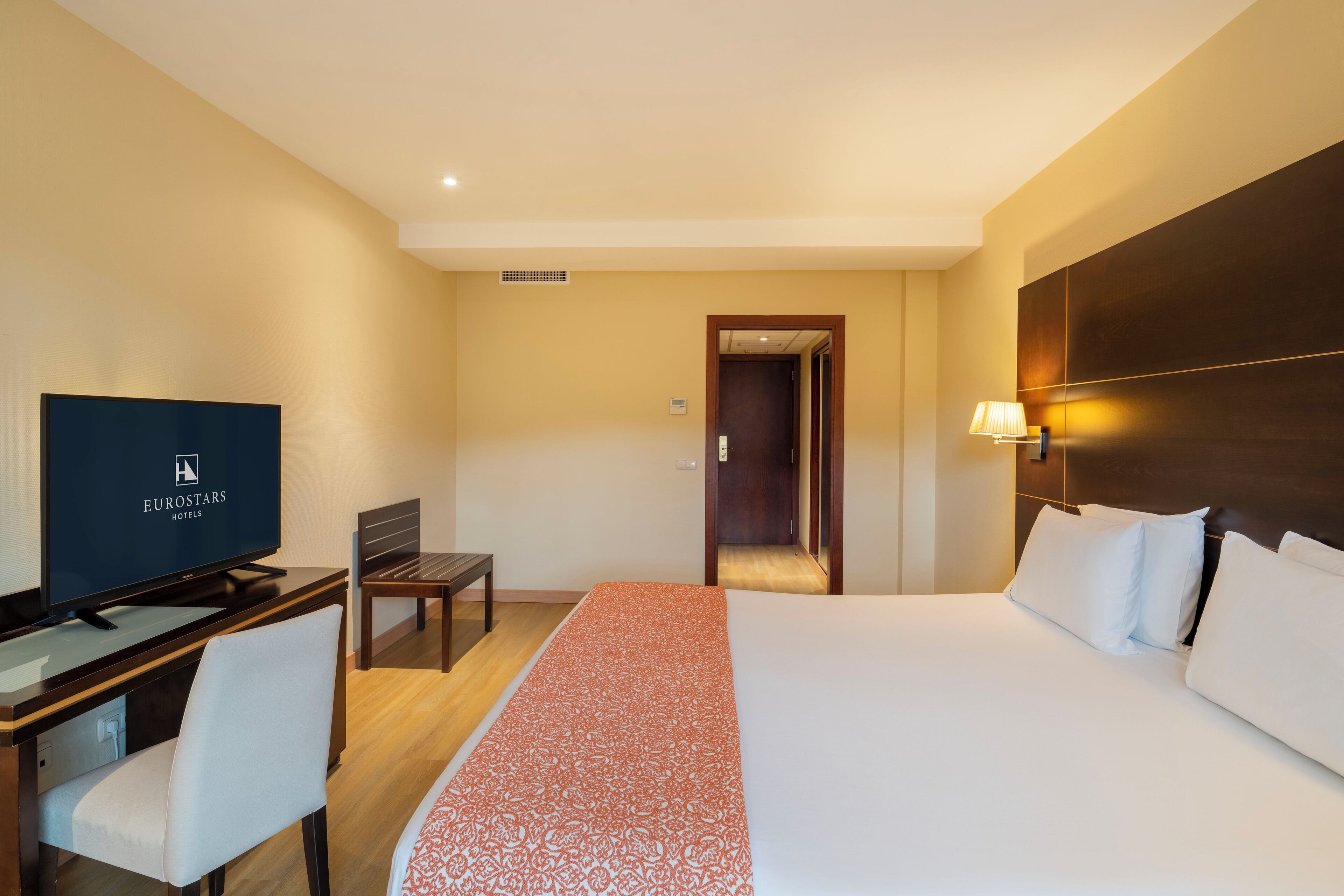 Una habitación de hotel con una cama, un escritorio con televisor, una silla y una mesita de noche.