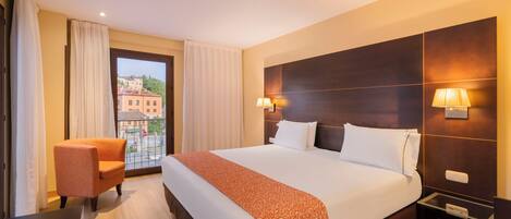Habitación de hotel con una cama grande, una silla, una mesita de noche y vistas a edificios a través del ventanal.