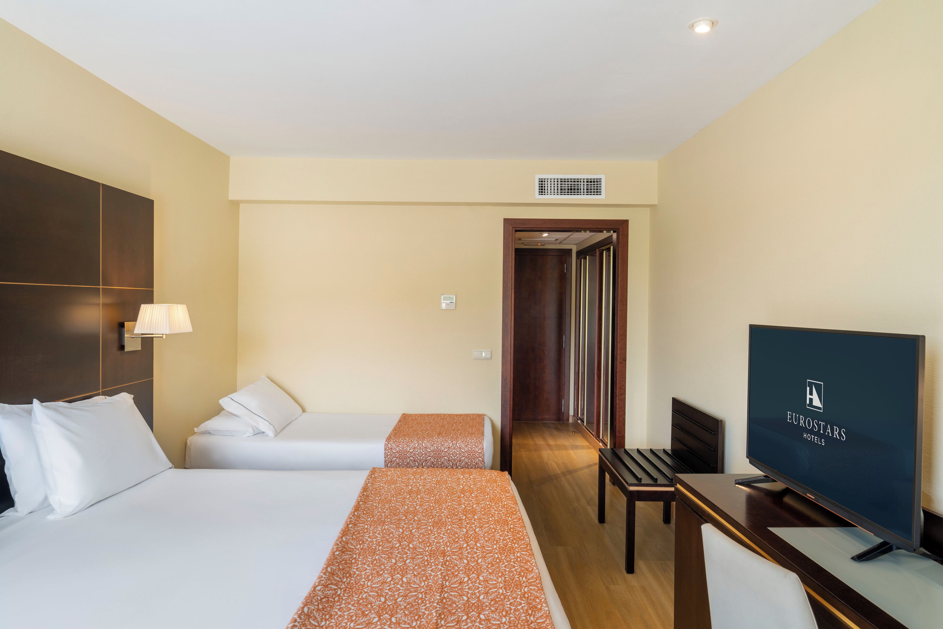 Quarto de hotel com duas camas, uma escrivaninha com televisão e uma porta que dá para um corredor.