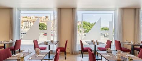 Un comedor de hotel con mesas y sillas, ventanas amplias con cortinas y vistas a edificios.