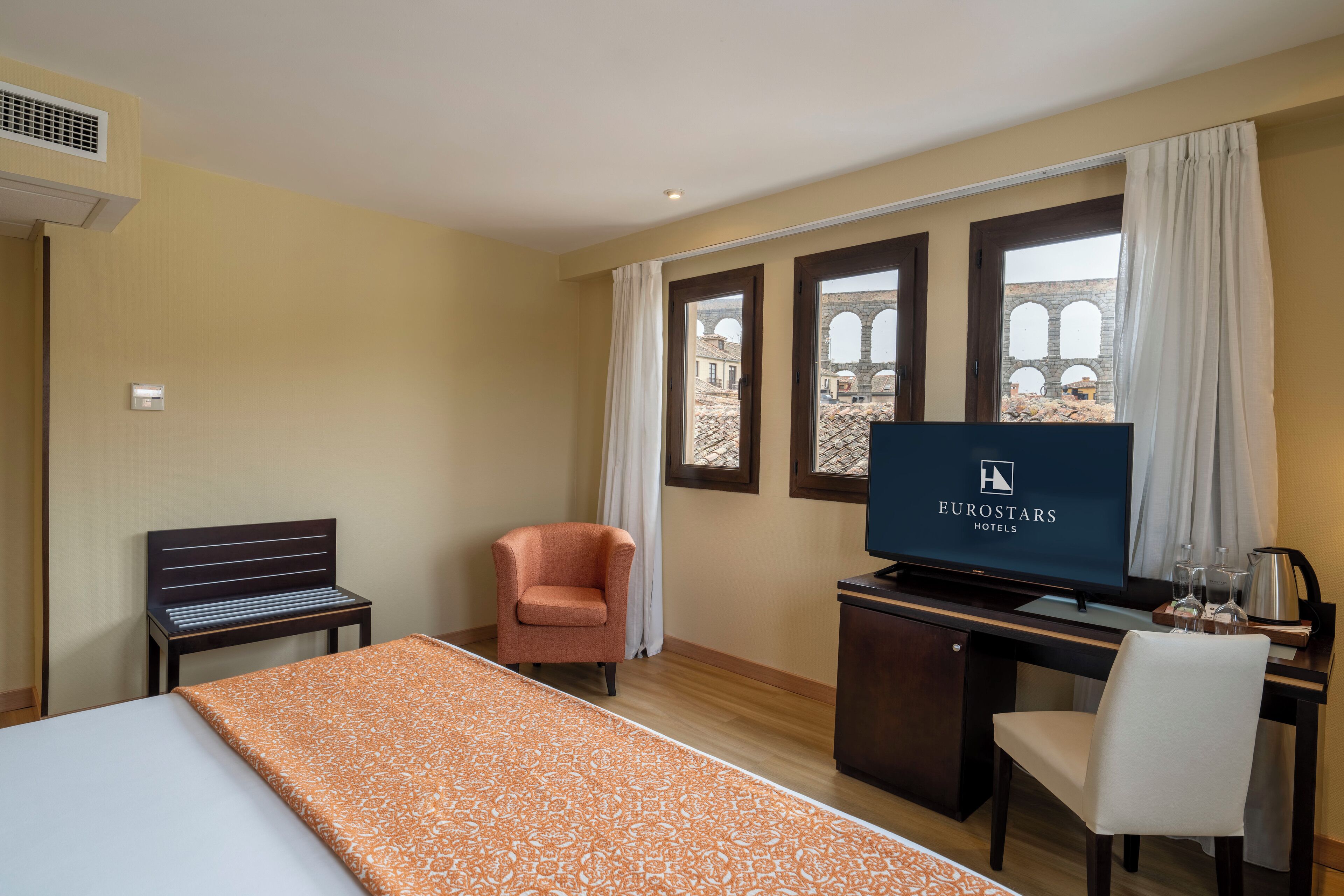Habitación de hotel con cama, escritorio con televisor, una silla y vista a una estructura antigua a través de las ventanas.