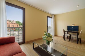 A hotel room with a sofa, a glass table with a vase, a TV, and a balcony with a view of buildings.