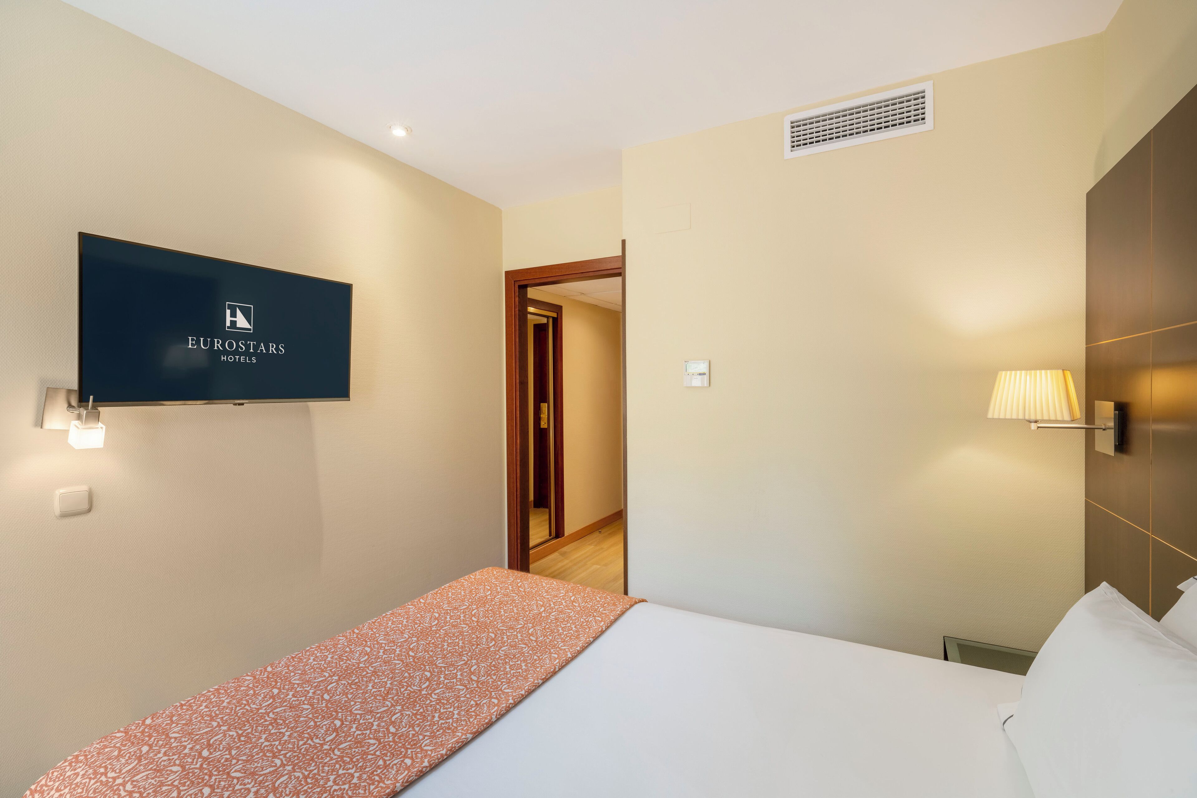 A hotel room with a bed, a television mounted on the wall, and a lamp.