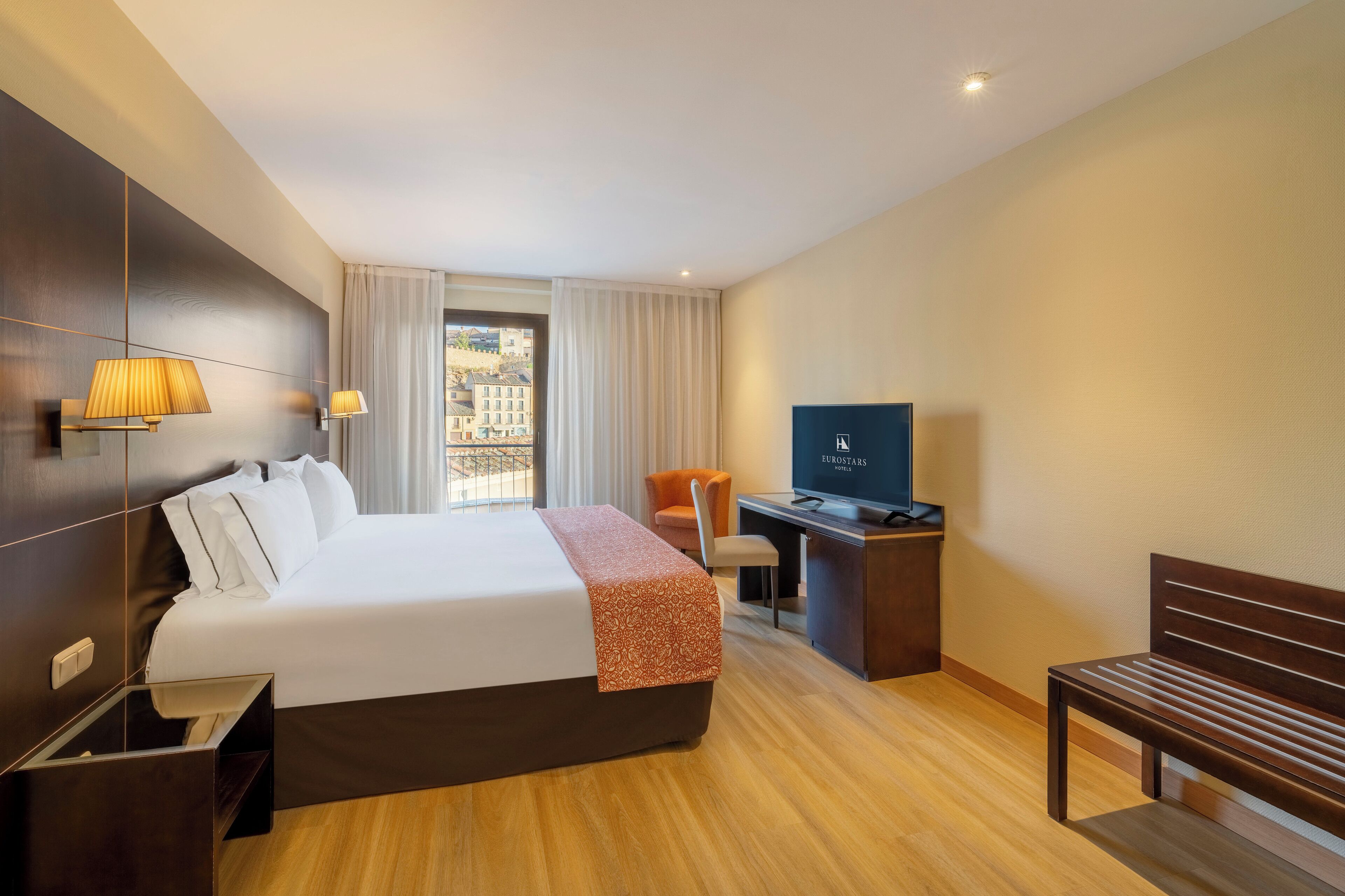 Una habitación de hotel con una cama grande, un escritorio con televisión, una silla y un banco.