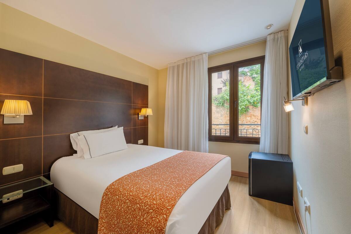 Hotelkamer met een groot bed, twee bedlampen, een aan de muur bevestigde televisie en een raam met gordijnen.