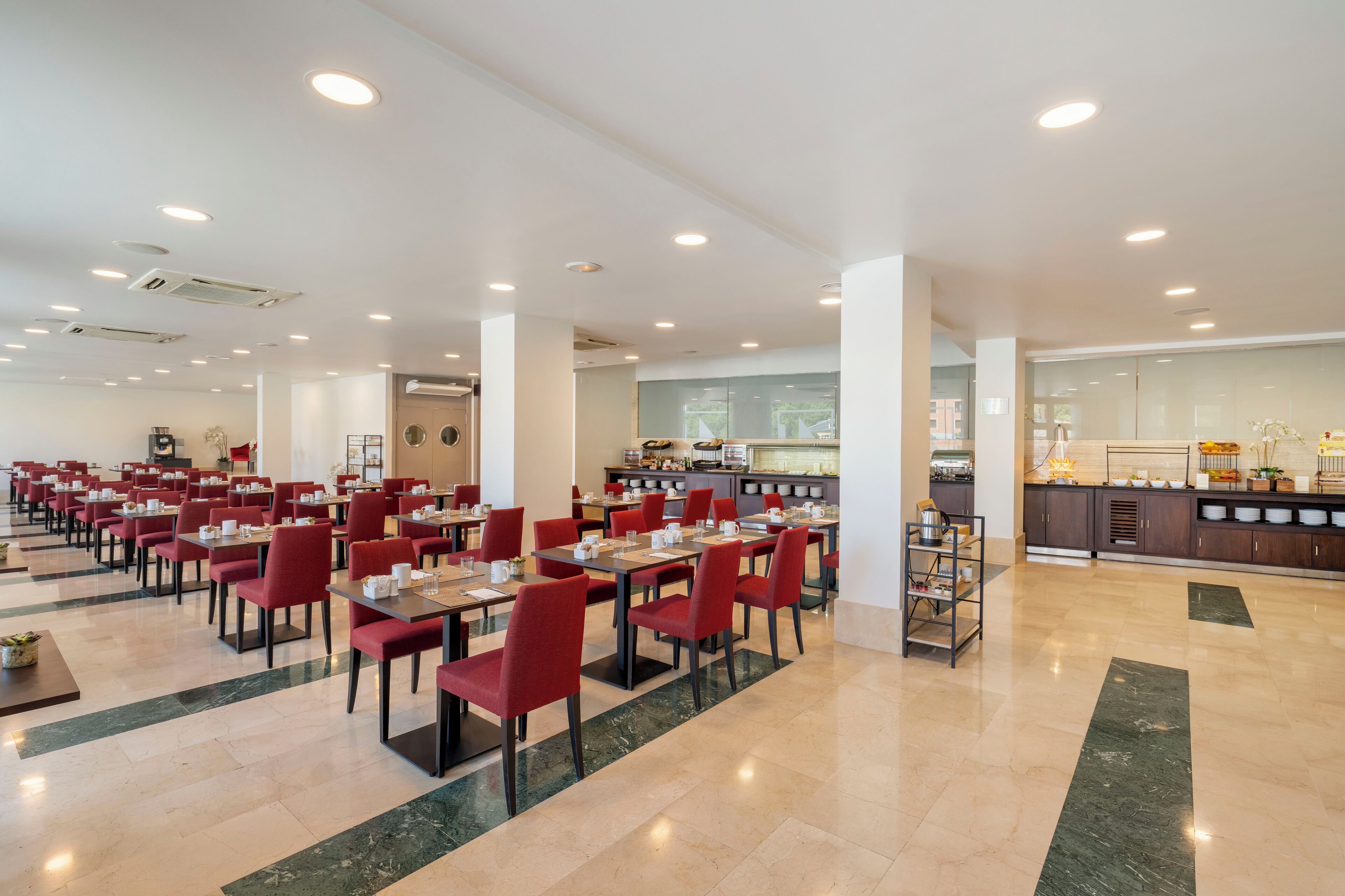 A spacious dining area with red chairs and tables, a kitchen in the background, and a clean, modern design.