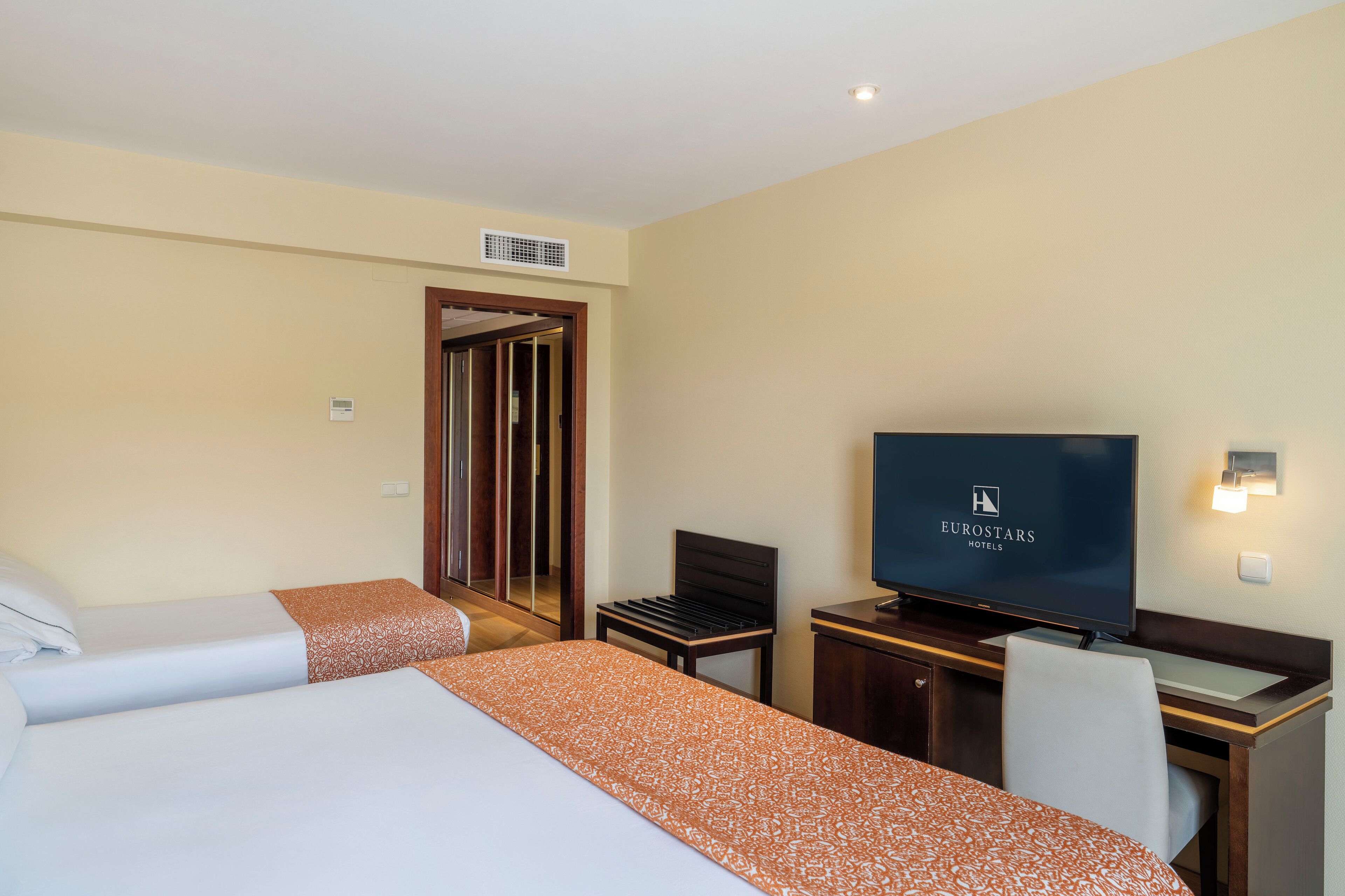 Habitación de hotel con dos camas, un escritorio y un televisor, y una puerta que da a un baño.