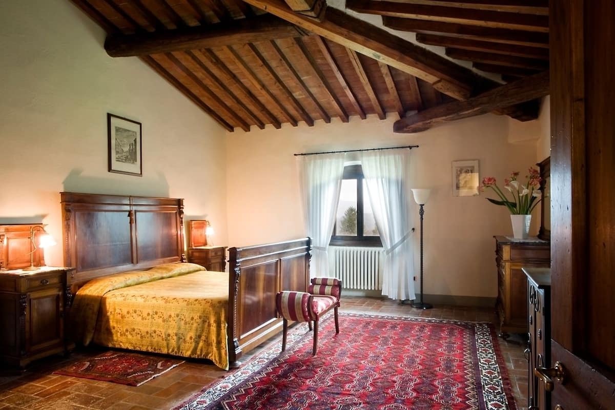 Un dormitorio con una cama de madera, un tapete rojo estampado, una silla, una ventana con cortinas y un radiador.