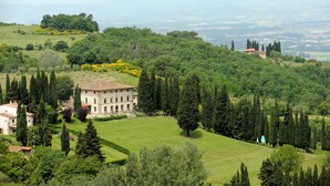 Un edificio grande e histórico rodeado de abundante vegetación y árboles, con un césped cuidado y una vista distante de montañas.